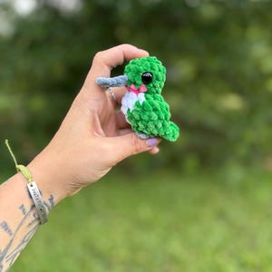 May include: A small, handmade crochet hummingbird plush toy featuring a vibrant green body, a white chest, a long grey beak, and a small pink accent under the chin. The bird has a single black plastic eye visible on the side of its head.
