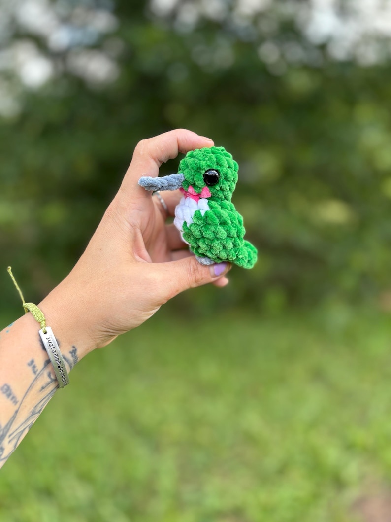 May include: A small, handmade crochet hummingbird plush toy featuring a vibrant green body, a white chest, a long grey beak, and a small pink accent under the chin. The bird has a single black plastic eye visible on the side of its head.