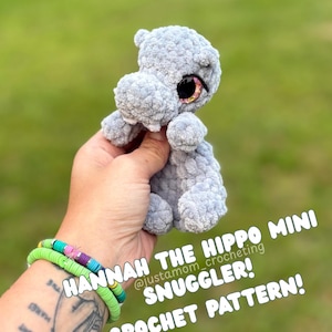 May include: A crocheted gray hippo mini snuggler with sparkly eyes. The text on the image reads "HANNAH THE HIPPO MINI SNUGGLER! CROCHET PATTERN!"