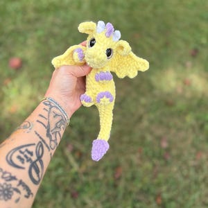 May include: A handmade yellow crochet dragon with purple accents on its feet, wings, and horns. The dragon has large black eyes and is held against a blurred green background. The dragon is a whimsical, soft toy.