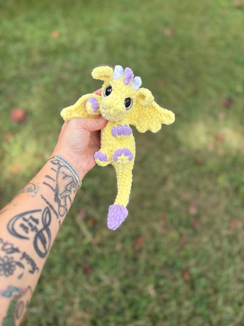 May include: A handmade yellow crochet dragon with purple accents on its feet, wings, and horns. The dragon has large black eyes and is held against a blurred green background. The dragon is a whimsical, soft toy.