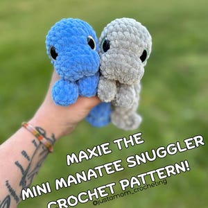 May include: Two crocheted manatee plushies, one blue and one gray, with large eyes. The plushies are being held in a hand. The text "Maxie the Mini Manatee Snuggler Crochet Pattern! @justamom_crocheting" is superimposed on the image.