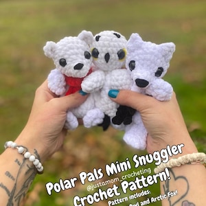 May include: A crochet pattern for Polar Pals Mini Snugglers, including a polar bear, snow owl, and arctic fox. The white stuffed animals have black eyes and noses. The polar bear has a red scarf. The text reads "Polar Pals Mini Snuggler Crochet Pattern!"