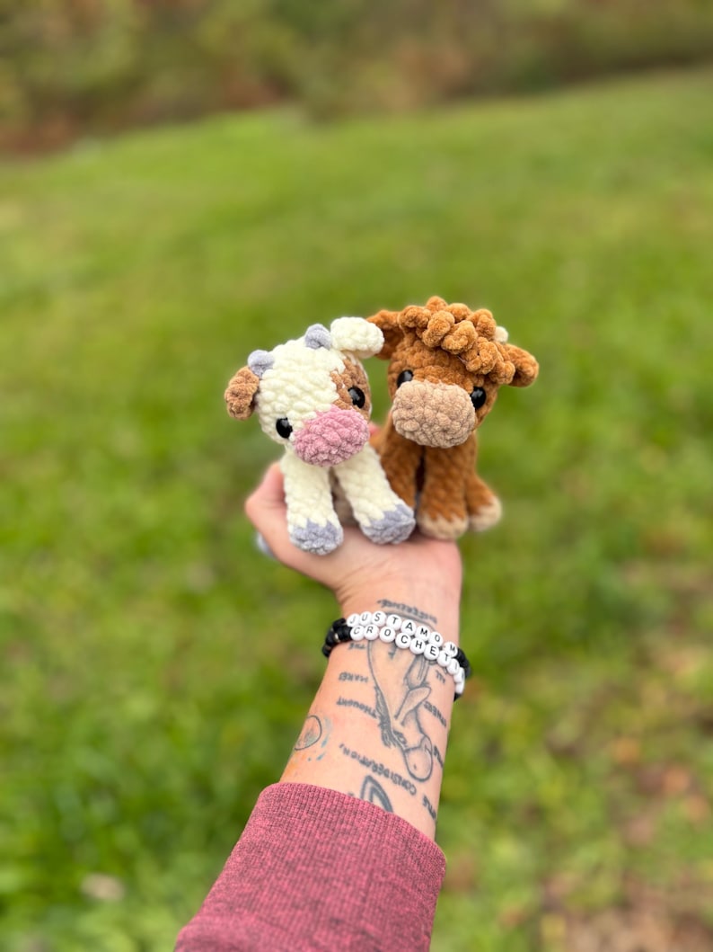 May include: Two handmade crochet cow plushies. One is white and brown with a pink nose, the other is brown. The plushies are held up against a blurred green background.