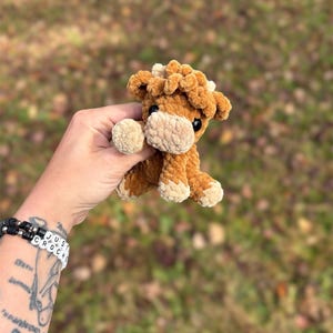 May include: A small, handmade crochet cow plushie in shades of brown and tan. The plush toy has a fluffy texture, with a light tan muzzle, hooves, and inner ears. The cow has small black eyes and is held against a blurred outdoor background.