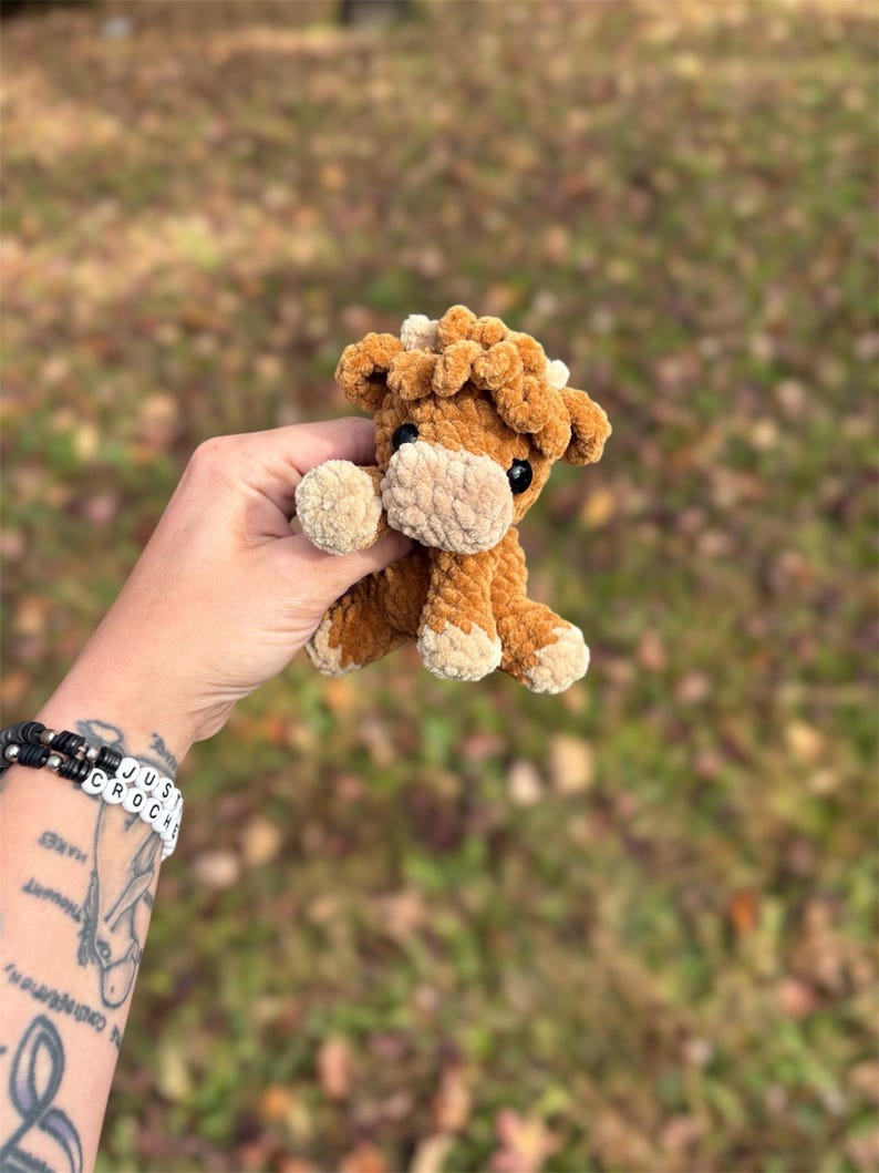 May include: A small, handmade crochet cow plushie in shades of brown and tan. The plush toy has a fluffy texture, with a light tan muzzle, hooves, and inner ears. The cow has small black eyes and is held against a blurred outdoor background.