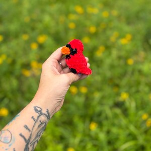 May include: A small, handcrafted plush cardinal bird made of soft red yarn. The bird features a bright orange beak, black eye patches, and a small black detail on its chest. The texture of the yarn is fuzzy and dense, creating a rounded, compact shape.