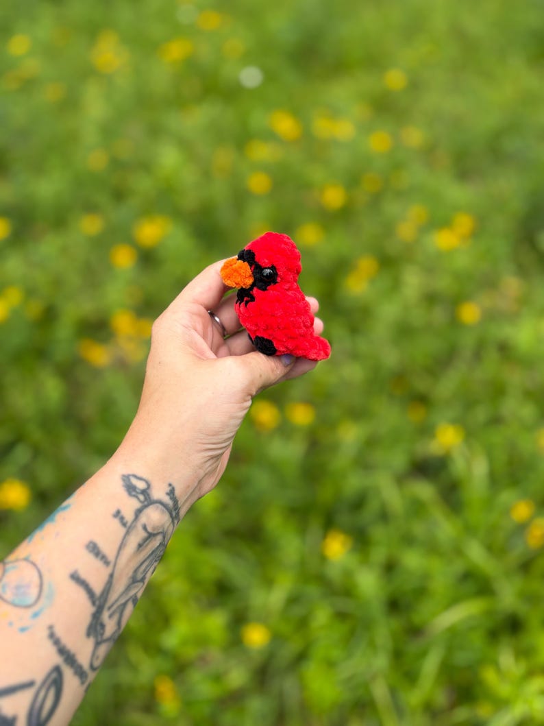 May include: A small, handcrafted plush cardinal bird made of soft red yarn. The bird features a bright orange beak, black eye patches, and a small black detail on its chest. The texture of the yarn is fuzzy and dense, creating a rounded, compact shape.
