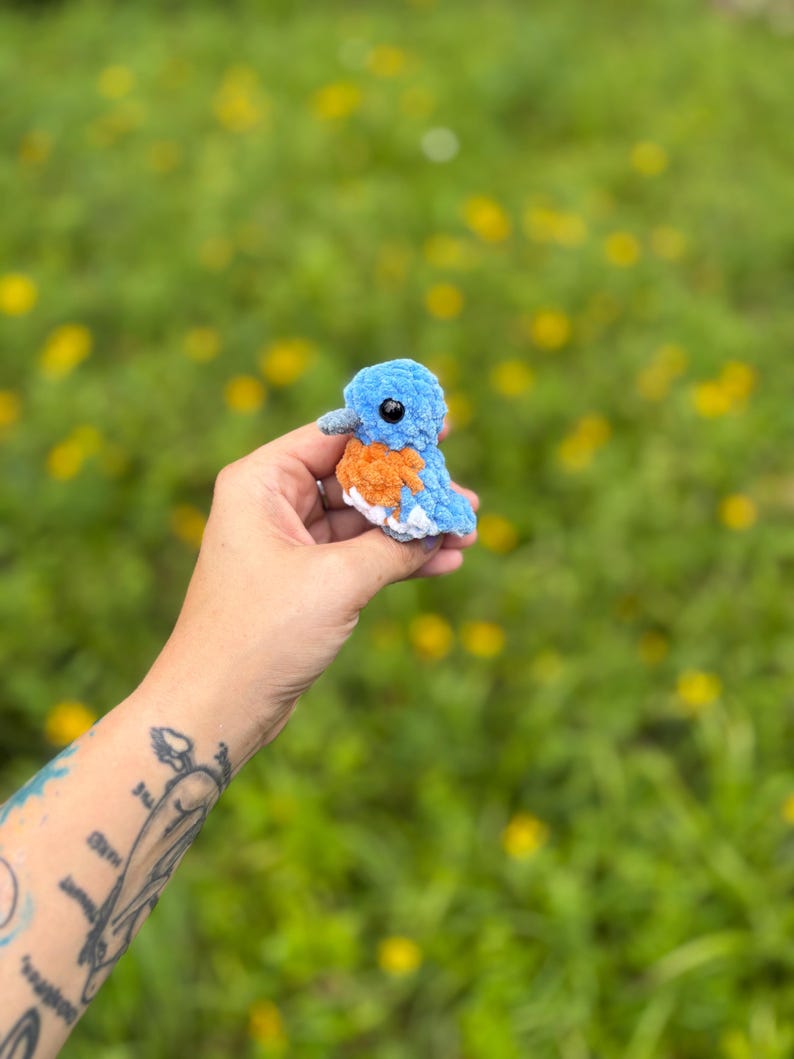 May include: A small, handmade crochet bird plushie featuring a light blue head and back, an orange chest, and white underparts. The bird has a small grey beak and round black plastic eyes, crafted from soft, textured yarn in a compact size.