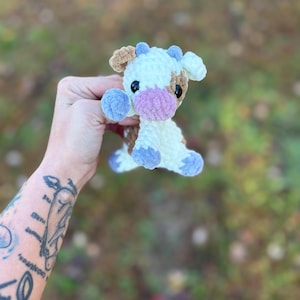 May include: A handmade crochet cow plushie in white, brown, and blue. The cow has a pink muzzle, black eyes, and blue accents on its hooves and horns. The plushie is soft and cuddly, perfect for a child's toy or nursery decor.