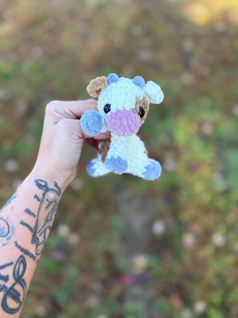 May include: A handmade crochet cow plushie in white, brown, and blue. The cow has a pink muzzle, black eyes, and blue accents on its hooves and horns. The plushie is soft and cuddly, perfect for a child's toy or nursery decor.