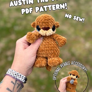 May include: A crocheted brown otter plushie with blue eyes and a light brown muzzle. The text "Austin the Otter PDF Pattern! No Sew!" is displayed above the plushie. An optional pebble accessory is shown in a smaller image.