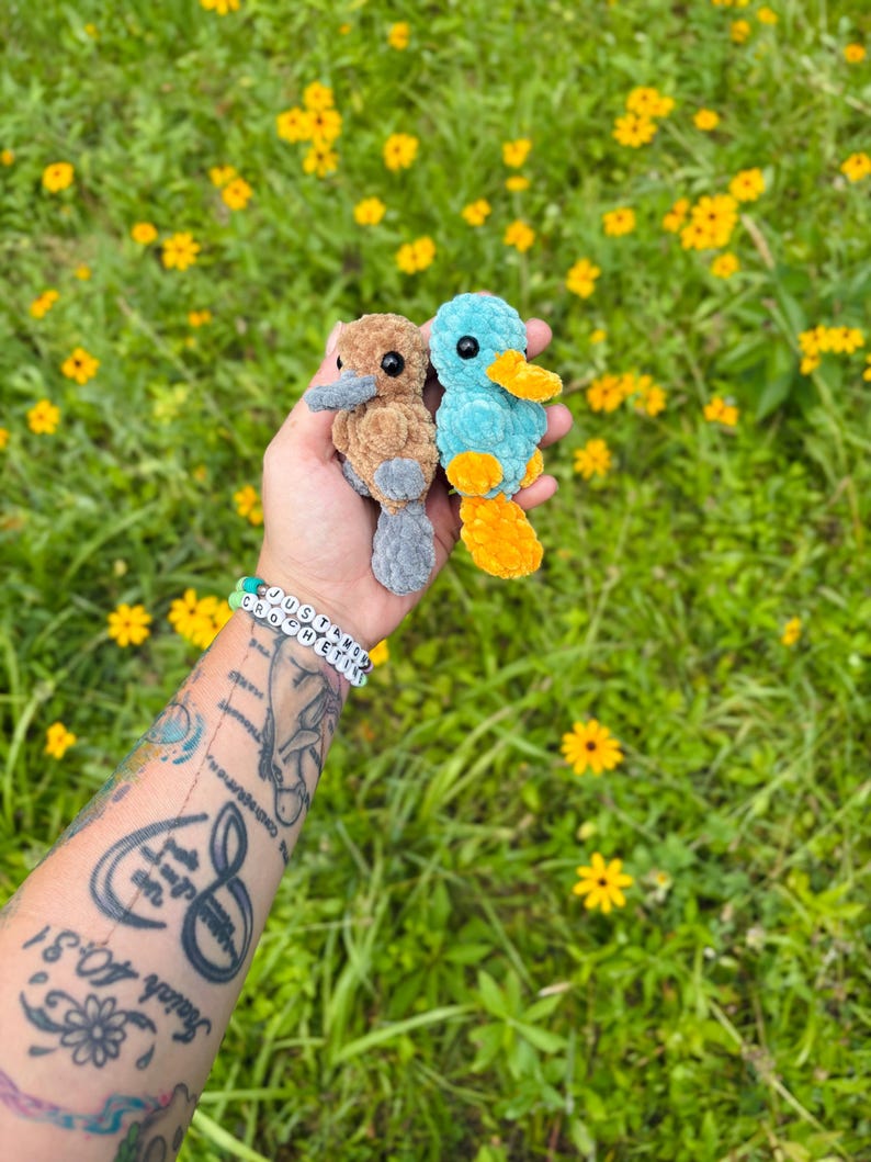 May include: Two handmade crochet platypus toys, one brown and gray, the other blue and yellow, are held in front of a background of green grass and yellow flowers. The toys have black eyes and are made of soft yarn.