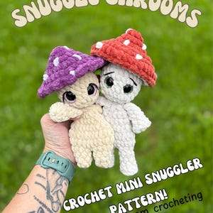 May include: Two crochet mushroom plushies, one beige and one white, with mushroom caps in purple and red, each with white polka dots. The text "SNUGGLE SHROOMS" is arched above the mushrooms. The text "CROCHET MINI SNUGGLER PATTERN!" and "@justamom_crocheting" are below.