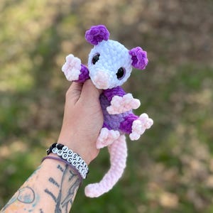 May include: A handmade crochet opossum plushie in shades of purple, white, and pink. The plush toy has a light purple body, white head, and pink nose and feet. The opossum has black eyes and a long, pink tail. The plushie is approximately 15 cm tall.