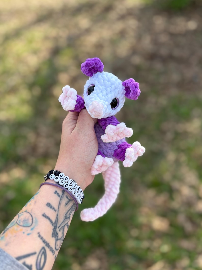 May include: A handmade crochet opossum plushie in shades of purple, white, and pink. The plush toy has a light purple body, white head, and pink nose and feet. The opossum has black eyes and a long, pink tail. The plushie is approximately 15 cm tall.