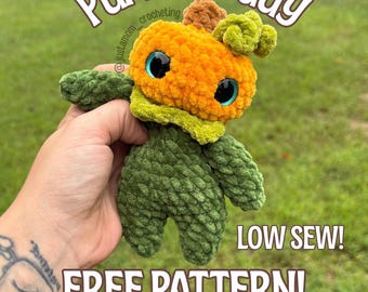 May include: A crocheted pumpkin figure with a bright orange head, a small brown stem, and a curly green leaf. The body is dark green with textured yarn. It features large, glossy blue eyes and text that reads BY JUST A MOM CROCHETING PUMPKIN GUY LOW SEW! FREE PATTERN! SAVE ME FOR LATER!