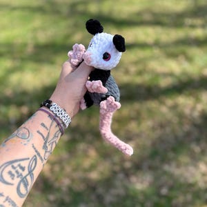 May include: A handmade crochet opossum plush toy. The opossum has a white head, black ears, and a grey body. It features pink paws and a pink tail. The toy is held against a blurred green background.