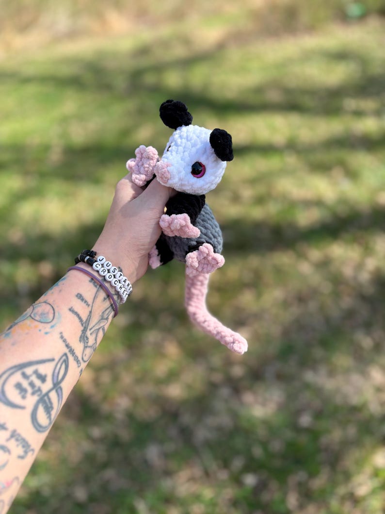 May include: A handmade crochet opossum plush toy. The opossum has a white head, black ears, and a grey body. It features pink paws and a pink tail. The toy is held against a blurred green background.