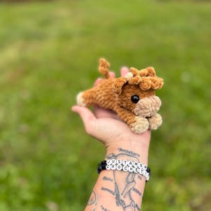 May include: A handmade, brown crochet cow plushie with a cream-colored muzzle and hooves. The cow has black eyes and a fluffy mane. The plushie is lying down, and is being held in a hand. A bracelet on the wrist reads "JUSTAMOM CROCHETI".