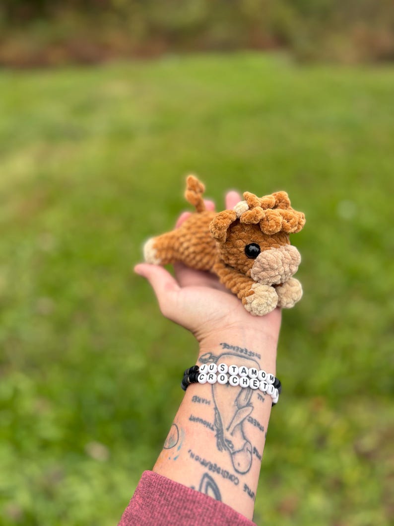 May include: A handmade, brown crochet cow plushie with a cream-colored muzzle and hooves. The cow has black eyes and a fluffy mane. The plushie is lying down, and is being held in a hand. A bracelet on the wrist reads "JUSTAMOM CROCHETI".