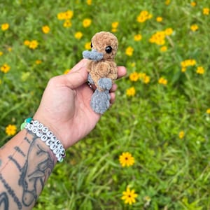 May include: A small, handmade crochet platypus in shades of brown and gray. The plush toy has black eyes and a gray bill and feet. The background is a field of green grass and yellow flowers.