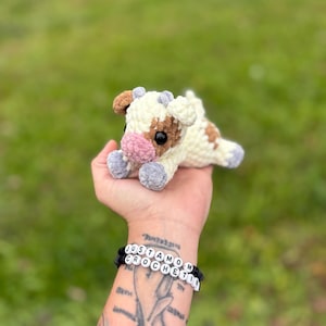 May include: A handmade crochet cow plushie in cream, brown, and pink. The cow has a soft, textured appearance and is lying down. The plushie is held in a hand, with two bracelets that read "JUSTAMOM CROCHET" in white letter beads.