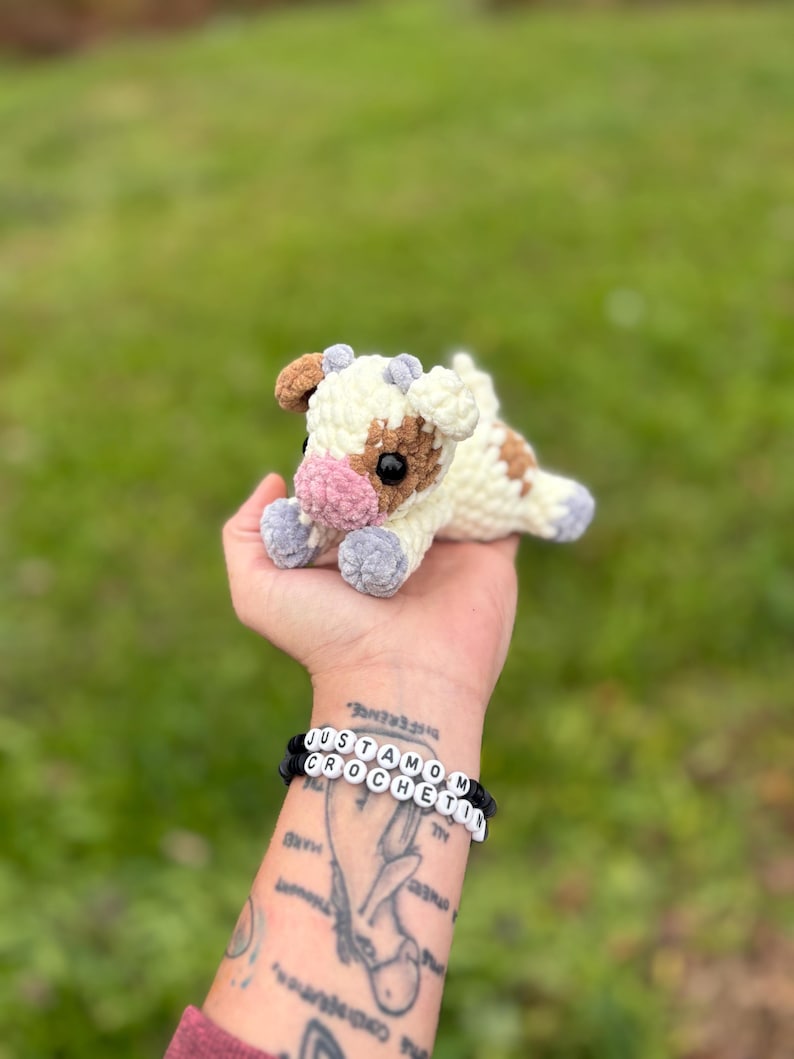 May include: A handmade crochet cow plushie in cream, brown, and pink. The cow has a soft, textured appearance and is lying down. The plushie is held in a hand, with two bracelets that read "JUSTAMOM CROCHET" in white letter beads.