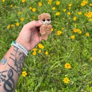 May include: A small, handmade crochet otter in shades of brown, held against a backdrop of green grass and yellow wildflowers. The otter has black eyes and a brown nose. A bracelet with the text "JUST MY CROCHET" is visible.