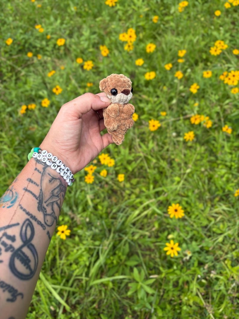 May include: A small, handmade crochet otter in shades of brown, held against a backdrop of green grass and yellow wildflowers. The otter has black eyes and a brown nose. A bracelet with the text "JUST MY CROCHET" is visible.