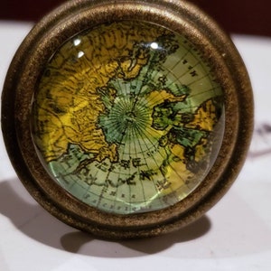 May include: A vintage-style brass knob with a glass globe featuring a world map. The map is in shades of green, yellow, and brown, with a compass rose in the center.