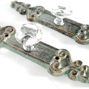 May include: Two ornate, distressed-finish cabinet pulls. Each has a rectangular base in teal and gold with a clear, faceted crystal knob. The decorative hardware is designed for furniture and home decor.