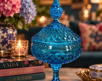 May include: A vibrant blue glass candy dish featuring a diamond-cut pattern across the bowl and lid. The piece stands on a pedestal base and is topped with a pointed finial. The glass is translucent and reflects light across its faceted surface.