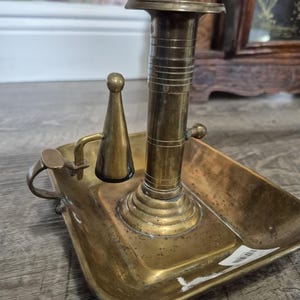 May include: A brass chamberstick candle holder featuring a rectangular tray base, a central cylindrical column with horizontal ribbed detailing, and an attached conical snuffer on a hinged arm with a small handle loop on the side.