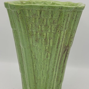 May include: A green ceramic planter with a textured, ribbed design. The planter has a square top and a flared, slightly curved shape. The surface has a speckled appearance.