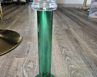 May include: A tall, slender candlestick holder featuring a deep green glass cylindrical stem and a circular base, topped with a clear, flared glass candle cup. The design is vertical and symmetrical, showcasing a smooth, polished glass finish.