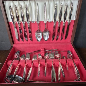 May include: A wooden silverware case with a red velvet interior, holding a complete set of silver-colored flatware. The set includes knives, forks, spoons, and serving utensils, all neatly arranged. The case is open, revealing the contents.