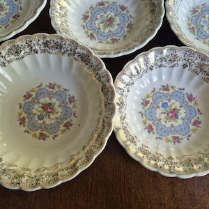 May include: A collection of vintage white ceramic bowls with a scalloped edge and gold trim. Each bowl features a central floral design in shades of blue, pink, and yellow. The bowls are arranged on a dark wooden surface.