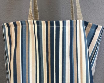 May include: A vertical striped tote bag with beige, white, and shades of blue stripes. The bag has beige straps and is made of fabric. The background is a solid gray color.