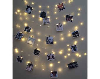 May include: A wall decorated with a string of fairy lights with photos clipped to them. The photos are of various sizes and subjects, including people, landscapes, and text. The lights are warm white and create a cozy atmosphere.