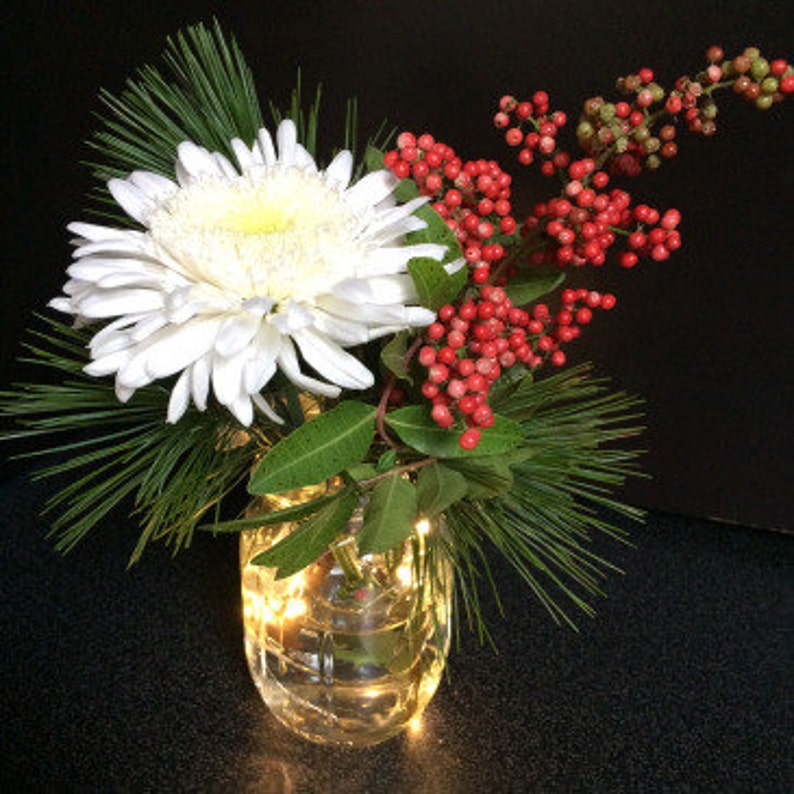May include: A white chrysanthemum flower, red berries, and green pine branches in a clear glass vase with white fairy lights.