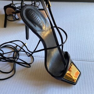 May include: Black leather heeled sandals with gold accents and lace-up detailing. The sandals have a square toe and a thin heel. The brand name "Schutz" is visible on the insole.