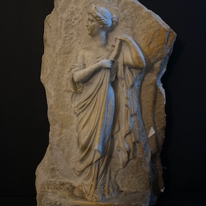 May include: A white plaster relief sculpture of a woman in a flowing robe, standing on a base with a Greek key pattern.