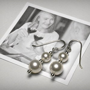 Peut inclure: Boucles d'oreilles pendantes en perles avec des détails argentés. Chaque boucle d'oreille comporte deux perles blanches rondes de tailles différentes, reliées par une petite perle argentée. Sur une photo en noir et blanc.