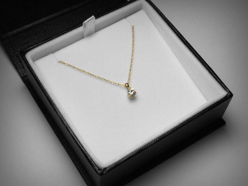 May include: A gold necklace with a single, round diamond pendant, presented in a black jewelry box with a white interior. The necklace chain is delicate, and the diamond sparkles.
