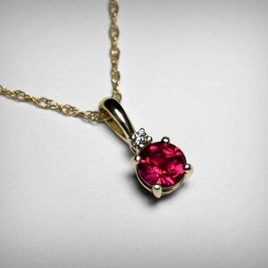 May include: A gold chain necklace with a pendant featuring a round, red gemstone surrounded by smaller, clear gemstones.