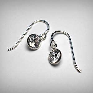 May include: A pair of silver-toned drop earrings. Each earring features a clear, round-cut gemstone set in a bezel setting. The earrings have a simple, curved hook design for pierced ears, and are displayed against a white background.