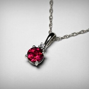 May include: A silver chain necklace with a pendant featuring a round, red gemstone and a smaller, clear gemstone.