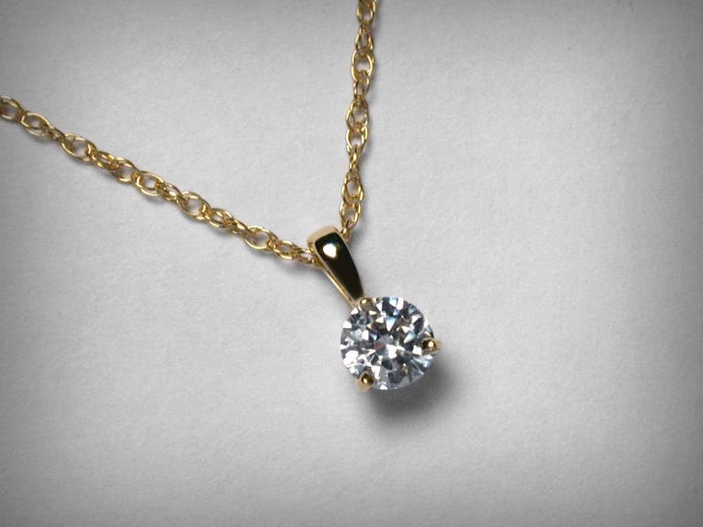 May include: A gold necklace with a round, clear diamond pendant. The diamond is set in a gold prong setting and hangs from a delicate gold chain. The jewelry is displayed on a light gray background.