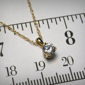 May include: A gold-colored necklace with a small, round, clear gemstone pendant. The chain is delicate and the pendant is set in a four-prong setting. The necklace is displayed against a white background with a ruler.